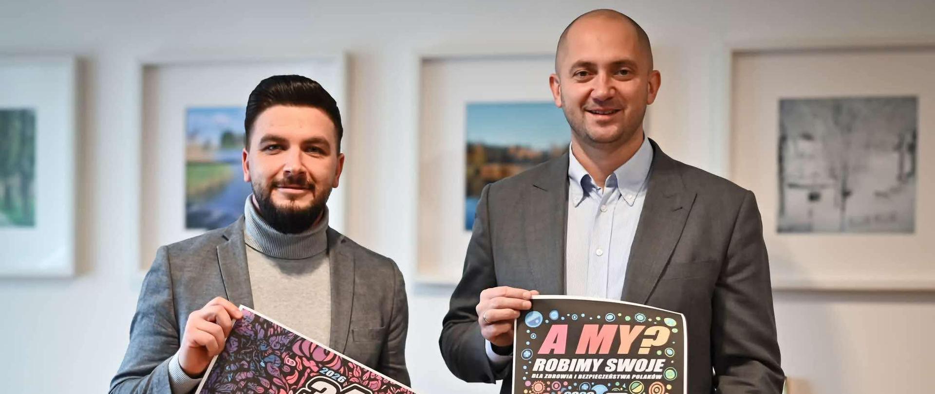 Dwóch mężczyzn w eleganckich marynarkach trzyma plakaty 34. Finału Wielkiej Orkiestry Świątecznej Pomocy w Konstantynowie Łódzkim. W tle wiszą obrazy na ścianie.