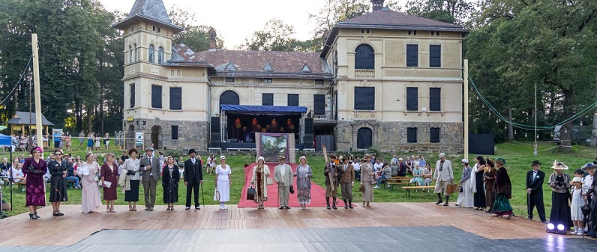 Uczestnicy wydarzenia stojący na scenie w strojach z końca XIX wieku. W tle pałac Głowińskich w Rabie Wyżnej