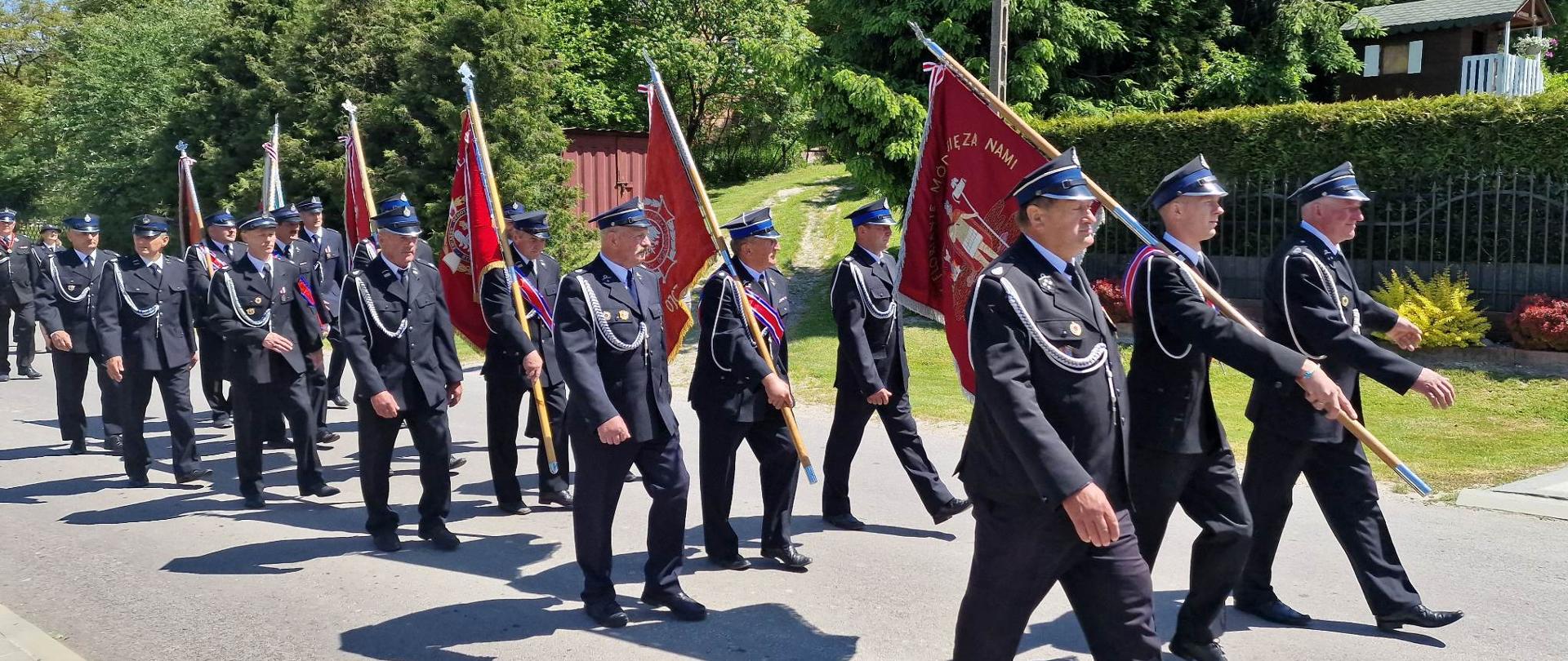 Zdjęcie przedstawia przemarsz pocztów sztandarowych jednostek Ochotniczych Straży Pożarnych. Strażacy w czarnych, galowych mundurach niosą sztandary swoich jednostek.