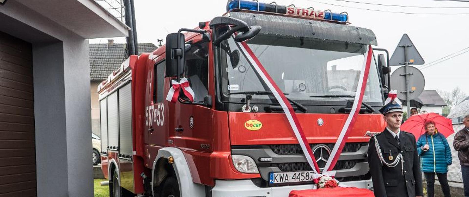 Czerwony wóz strażacki, na pierwszym planie stoi strażak ochotnik 