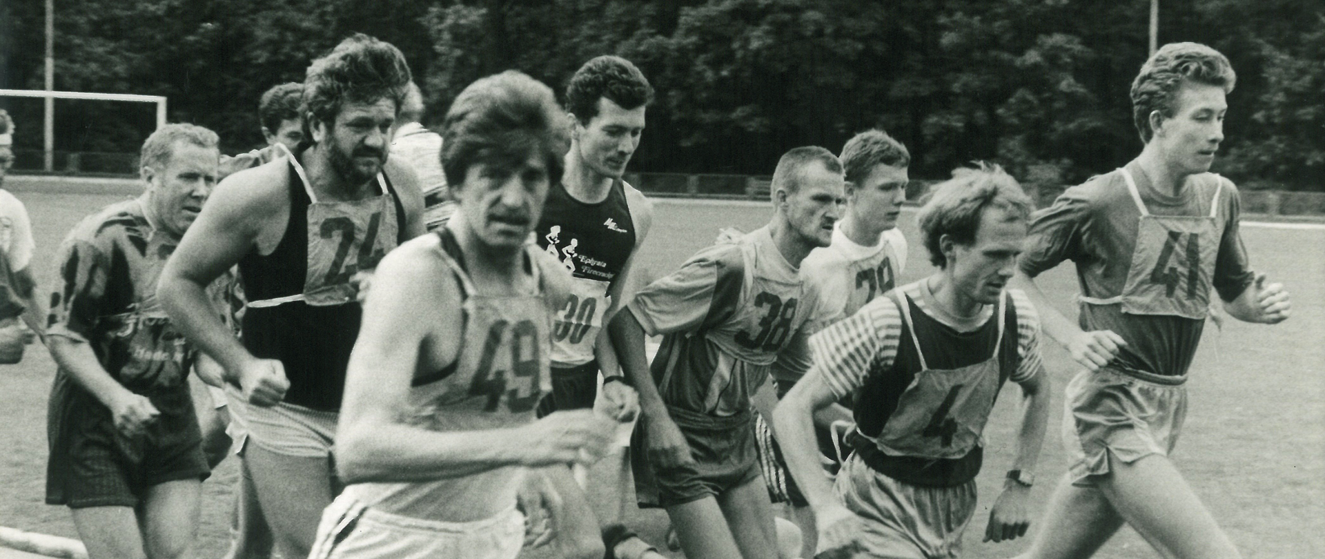 Grupa mężczyzn biegnie po bieżni podczas zawodów lekkoatletycznych. Uczestnicy mają numery startowe przypięte do koszulek. Zdjęcie w odcieniach szarości, wykonane najpewniej w latach 80. lub 90.