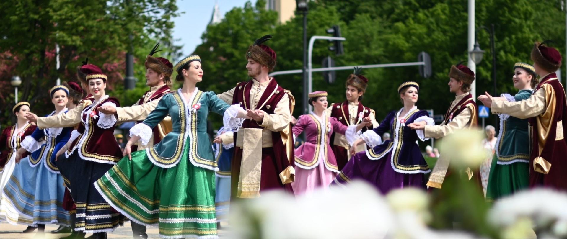 Zespół pieśni i tańca w tradycyjnych strojach z epoki tańczy poloneza. W tle wieża kościoła w Konstantynowie Łódzkim.