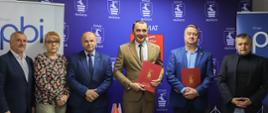 Pamiątkowe zdjęcie z podpisania umowy. Sześć osób stojących w rzędzie obok siebie na tle biało-niebieskich rollupów promocyjnych grupy PBI oraz niebieskiej ścianki Powiatu Kraśnickiego.
