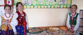 Stół regionalny z tradycyjnymi przysmakami, oscypki, korbace, moskole. Przy stole pozują dziewczynki w warkoczach i strojach góralskich.