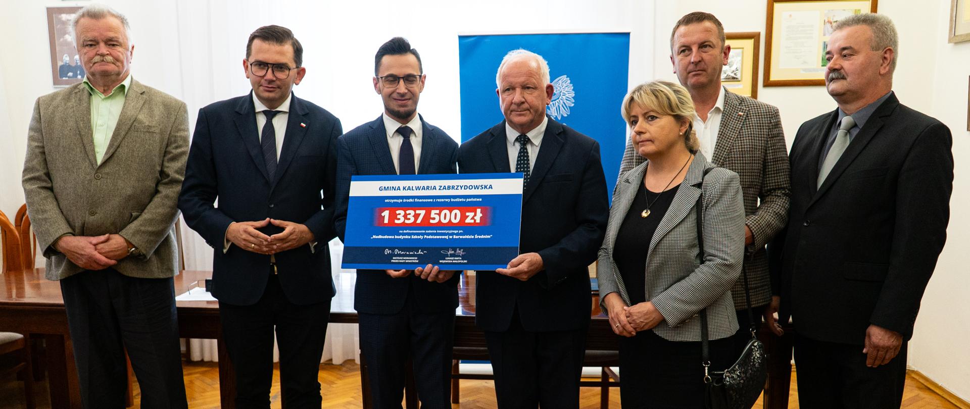 W gabinecie burmistrza pozują do fotografii grupowej uczestnicy spotkania. Stojący w środku poseł Kaczyński i burmistrz Ormanty trzymają symboliczny czek z kwotą dofinansowania.