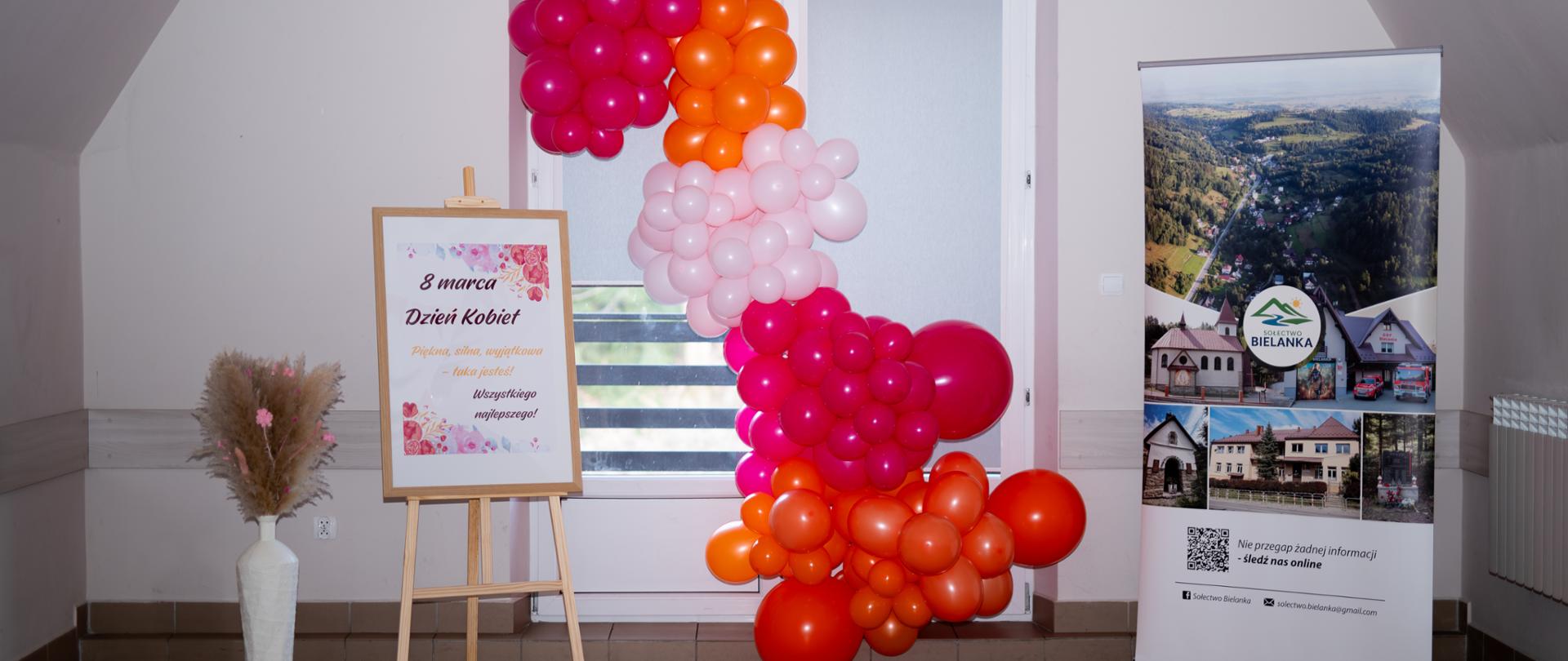 Po prawej baner sołectwa Bielanka, na środku ścianka z kolorowych balonów, po prawej sztaluga z plakatem informującym o dniu kobiet