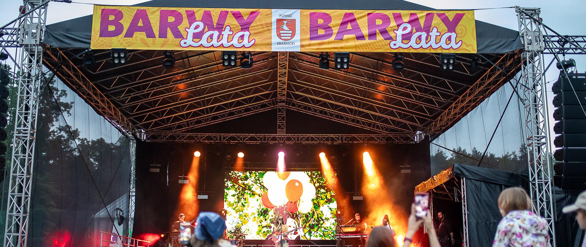 Widok na scenę plenerową z koncertu SARSA podczas wydarzenia „Barwy Lata” 2025 w Kraśniku. Na pierwszym planie licznie zgromadzona publiczność, na scenie artystka z zespołem w kolorowych światłach i wizualizacjach.