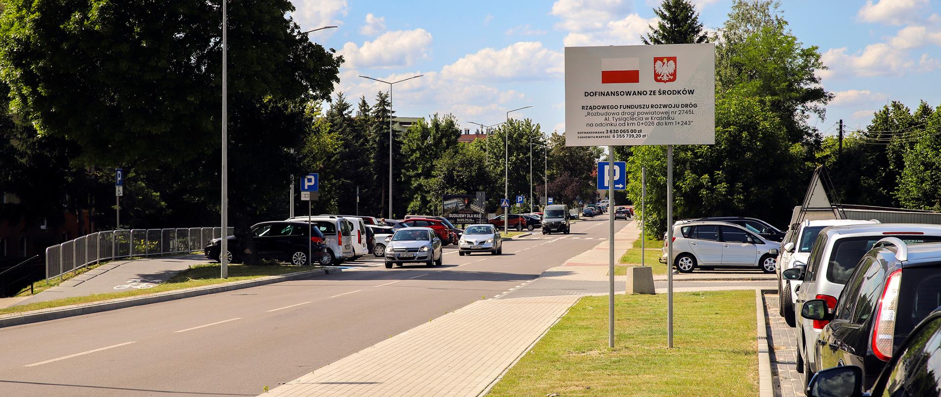 Na zdjęciu znajduje się droga asfaltowa, po której poruszają się auta. Przy drodze stoją zaparkowane samochody. Widać również wyremontowane chodniki. W pobliżu drogi znajduje się tablica informująca o dotacji otrzymanej na remont.