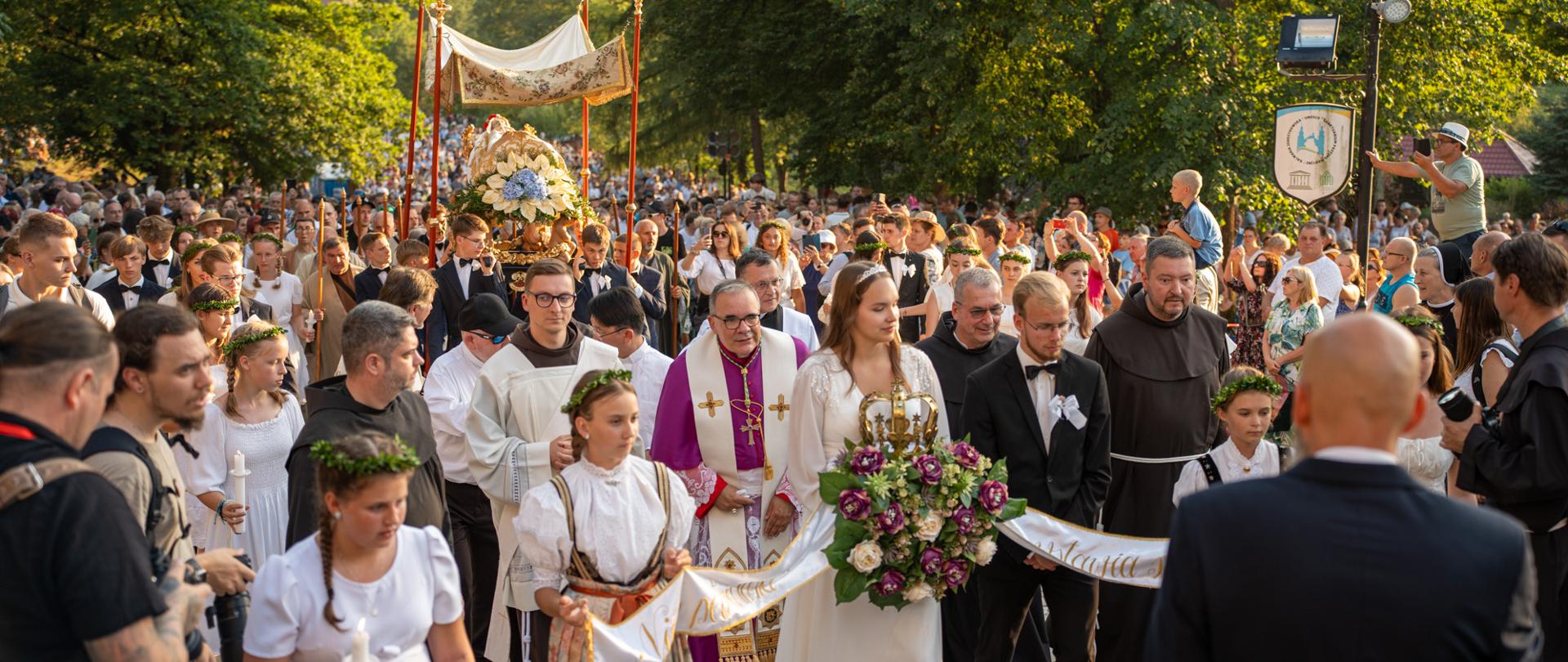 Uroczysta procesja religijna prowadzona przez kapłanów i osoby duchowne, z udziałem dziewcząt w białych sukienkach i tradycyjnych strojach ludowych, niosących świece i wstęgi. W centrum niesiona jest figura pod ozdobnym baldachimem, otoczona kwiatami. Wokół procesji zgromadził się tłum wiernych, a całość odbywa się w letniej scenerii, w otoczeniu zieleni i górskiego krajobrazu