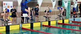 Pływalnia oraz widok na basen z wyznaczonymi torami oraz na zawodniczki, pływaczki stojące na słupkach w pozycji przygotowanej do skoku do wody. 