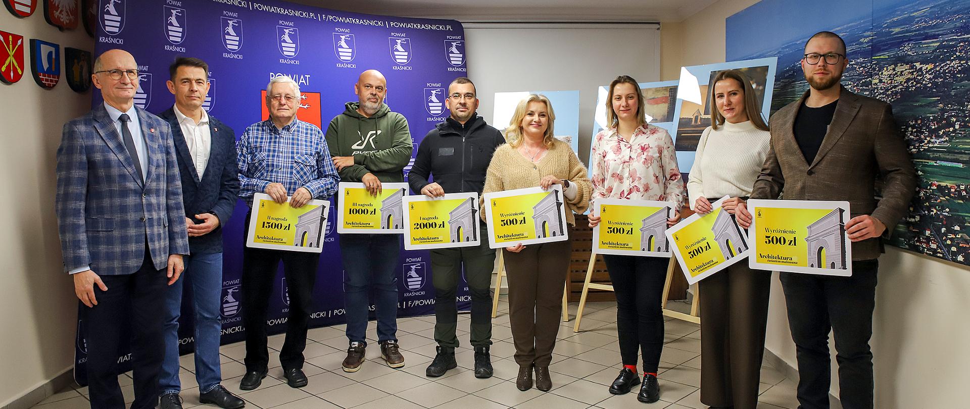 Zdjęcie przedstawia uczestników i organizatorów konkursu fotograficznego, stojących obok siebie w sali konferencyjnej w Starostwie Powiatowym w Kraśniku. W tle widoczna jest ścianka promocyjna Powiatu Kraśnickiego. Po prawej stronie znajdują się wydrukowane fotografie oprawione w ramy ustawione na sztalugach.
Uczestnicy trzymają w rękach duży, żółty bon podarunkowy z napisem, nazwą konkursu „Architektura światłem malowana” z kwotami nagrody. 