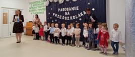 Przedszkolaki stoją w sali na tle ścianki "Pasowanie na przedszkolaka", obok nich nauczycielki, prowadzące uroczystość