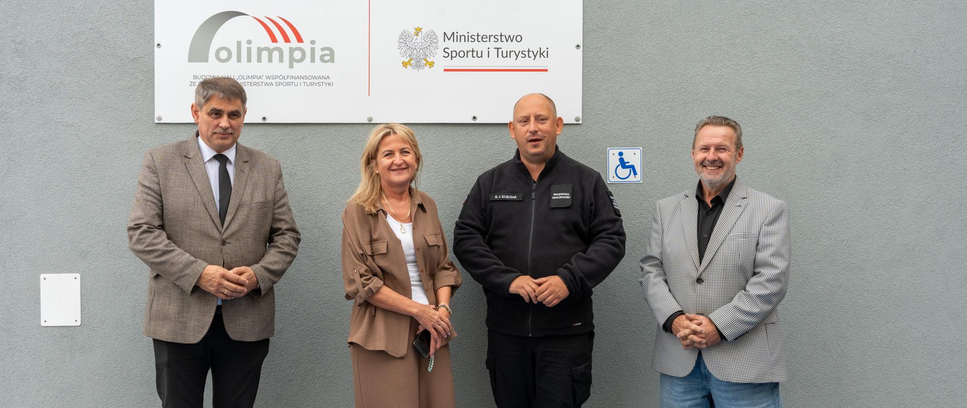 Cztery osoby, w tym wojewoda małopolski Krzysztof Jan Klęczar, pozują do wspólnego zdjęcia przed budynkiem szkoły w Zarzycach Wielkich, pod tablicą programu „Olimpia” Ministerstwa Sportu i Turystyki.