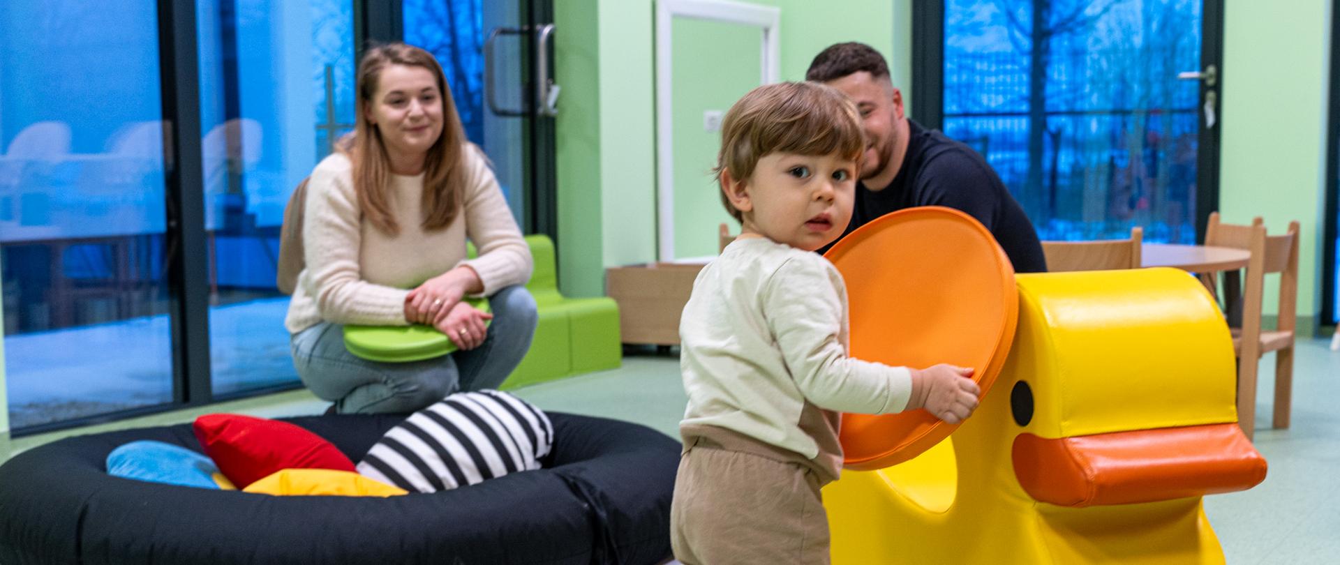 Małe dziecko bawi się kolorowym, żółtym elementem zabawowym w sali żłobka. W tle, na miękkich pufach i siedziskach, znajdują się rodzice obserwujący zabawę. Pomieszczenie jest jasne, nowoczesne i dostosowane do potrzeb najmłodszych dzieci.