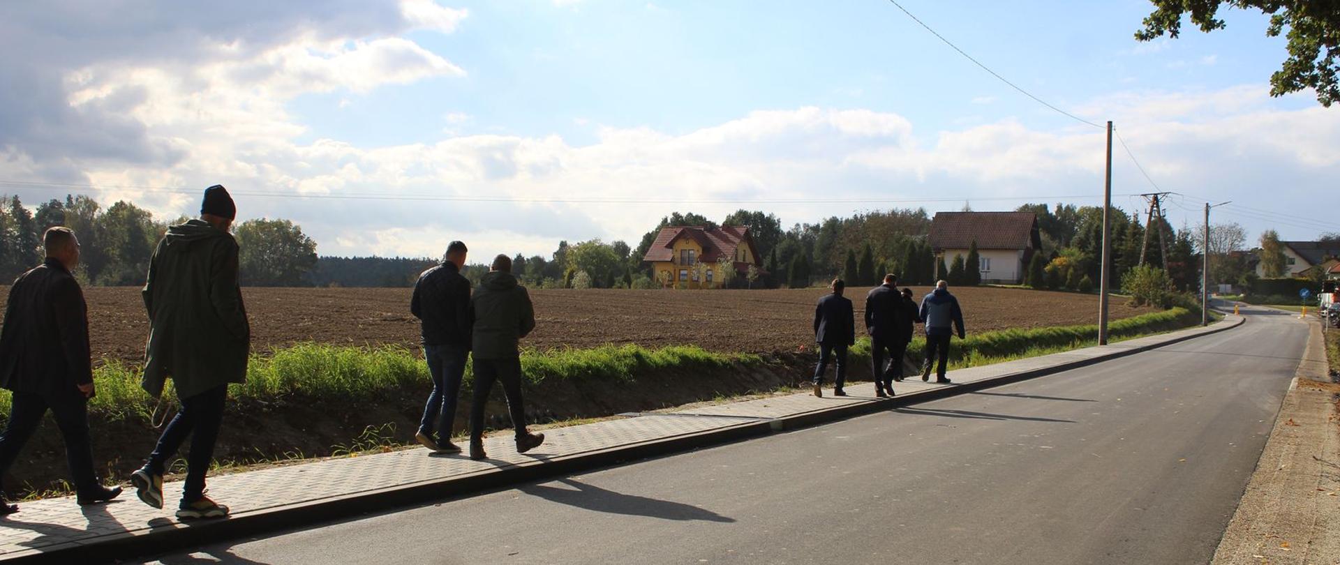 Droga gminna Wudyka w środkowej części zdjęcia widoczna droga wraz z chodnikiem i pieszymi, w tle zabudowania i pole rolne.