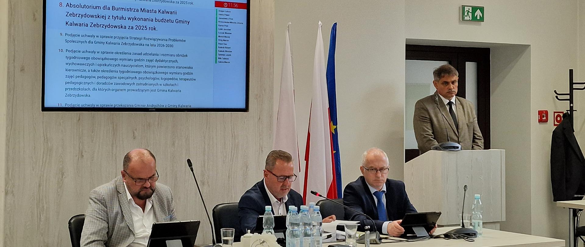 Na środku stół prezydialny Rady Miejskiej przy nim trzy osoby orezydium rady za nimi duzy monitor wyświetlający wyniki głosowania po prawej przy mównicy Burmistrz Miasta