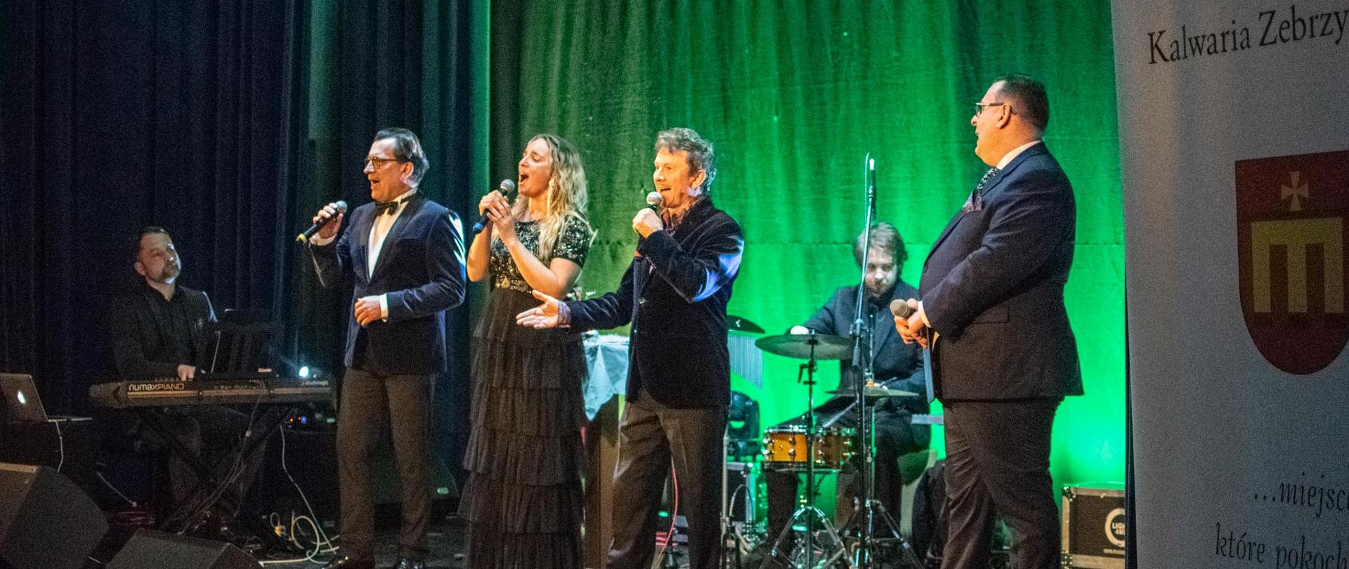 Na scenie znajduje się grupa artystów składająca się z 6 osób. Dwóch muzyków grających na instrumentach oraz trzech solistów znajdujących się w centrum zdjęcia. Obok nich stoi mężczyzna prowadzący koncert. 