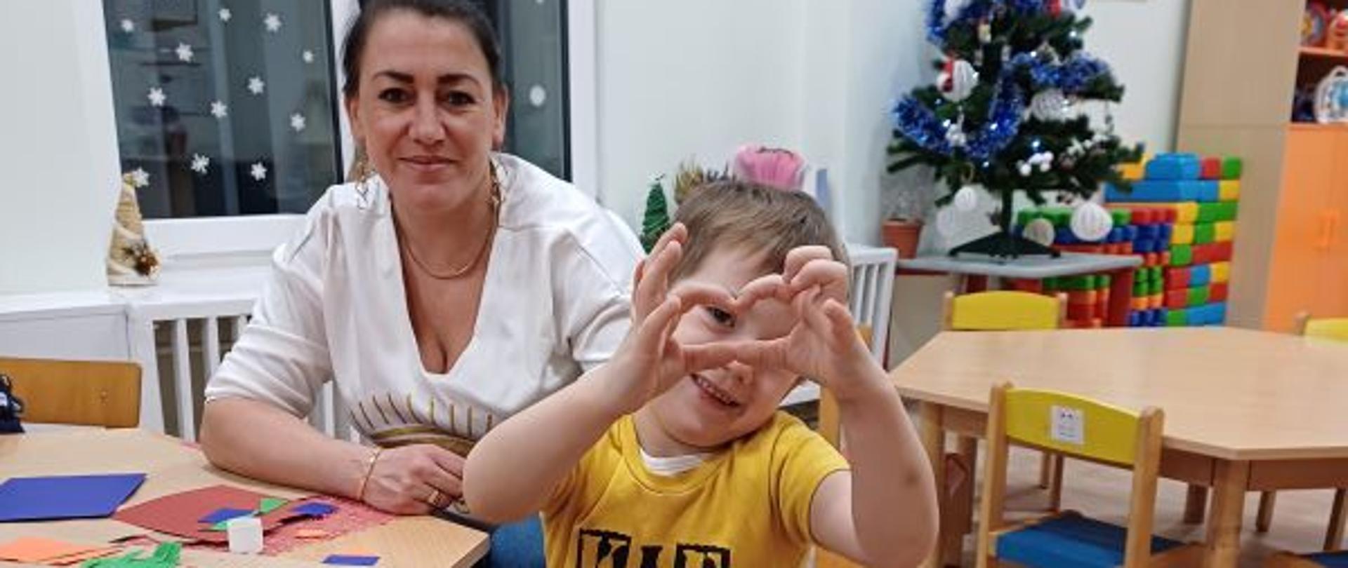 Chłopczyk pozuje do zdjęcia, pokazując serce splecionymi paluszkami dłoni, za nim siedzi dorosła kobieta
