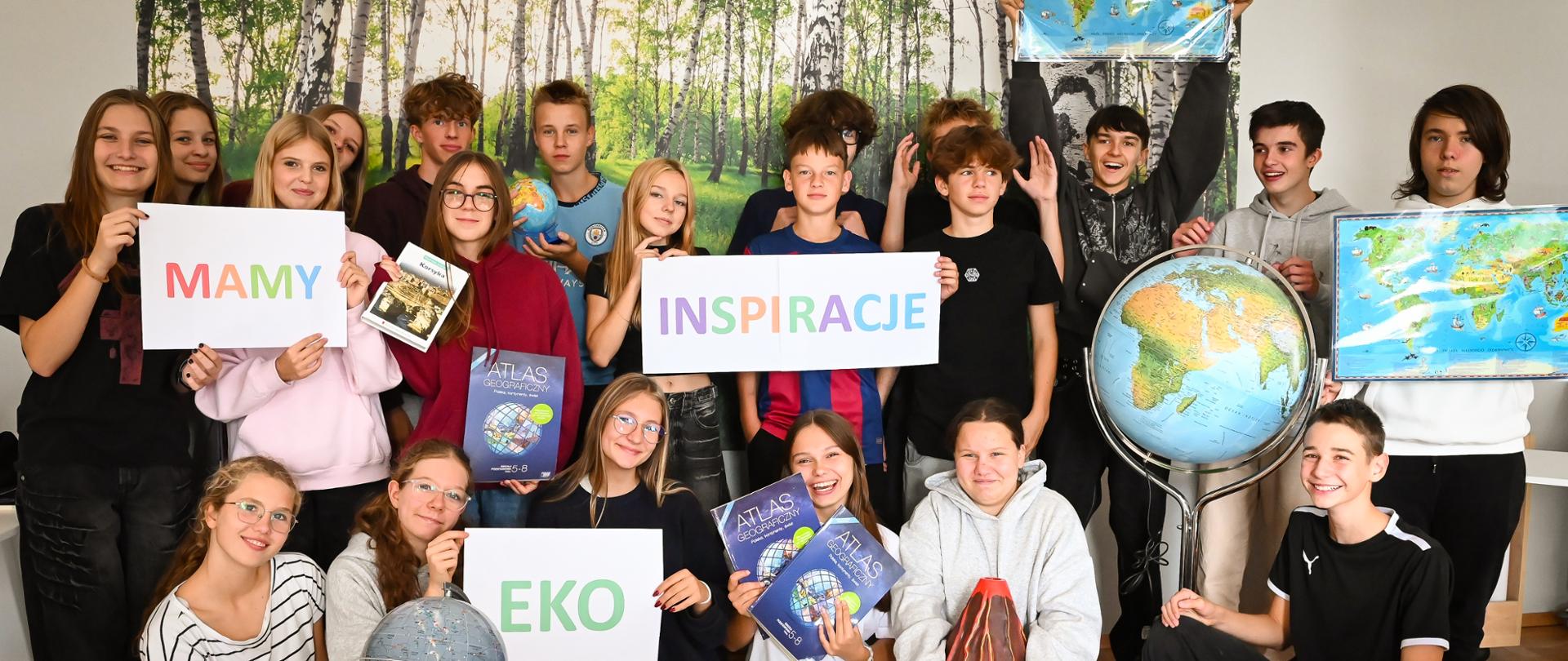 Grupa młodzieży pozuje wspólnie w sali lekcyjnej na tle ściany z fototapetą przedstawiającą brzozowy las. Uczniowie trzymają atlasy geograficzne, globusy i mapy, a także kartki z napisami: ‘MAMY’, ‘EKO’, ‘INSPIRACJE’. Na podłodze stoją modele edukacyjne, m.in. wulkan i układ planetarny.