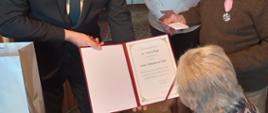 Moment wręczenia listu gratulacyjnego Państwu Halek przez Wójta Tomasza Rajcę. Wójt trzyma otwarty list go czyta a jubilaci patrzą w kierunku listu i słuchają. W tle w progu drzwi widać postać kobiety jest to chrześnica Pani Halek.