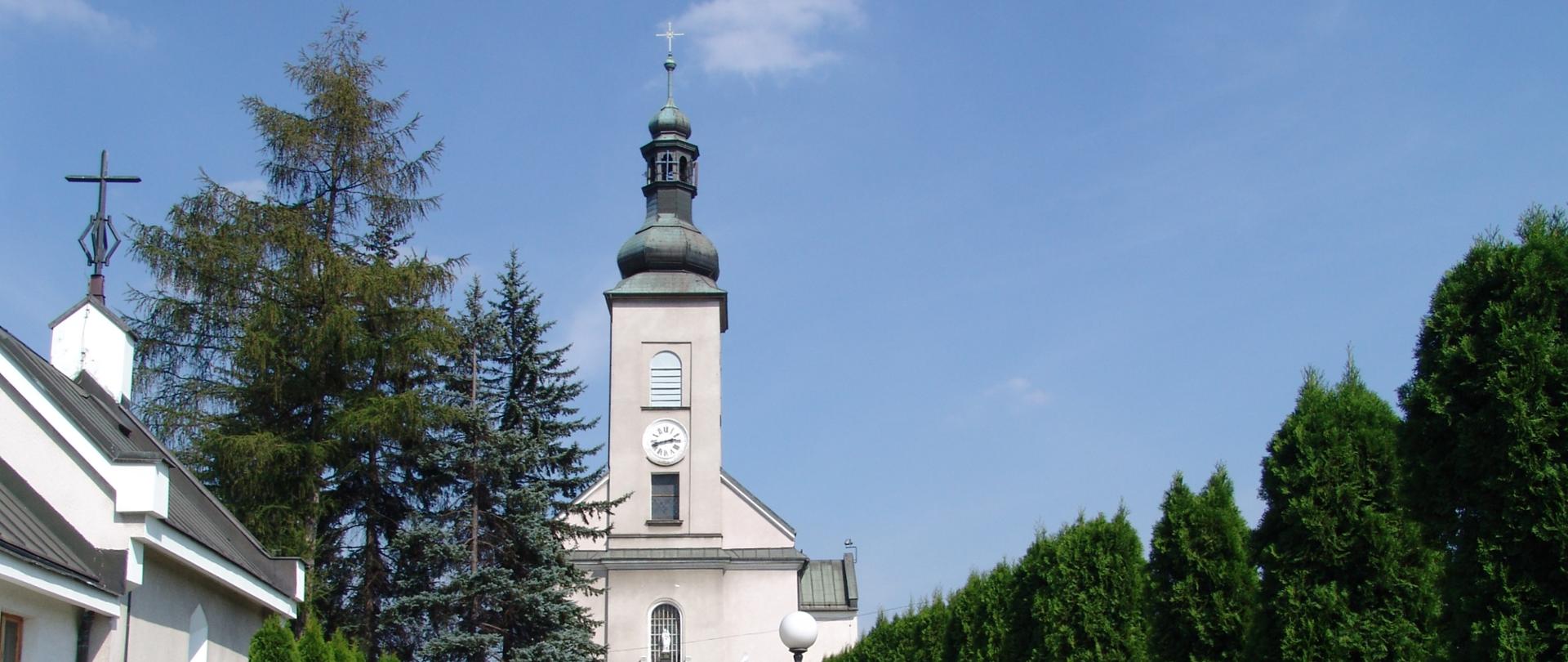 Widok na kościół z przykościelnego cmentarza. Po lewej stronie kaplica przedpogrzebowa, w środku brukowana alejka wiodąca w kierunku wejścia do kościoła. Po obu jej stronach rosną tuje.