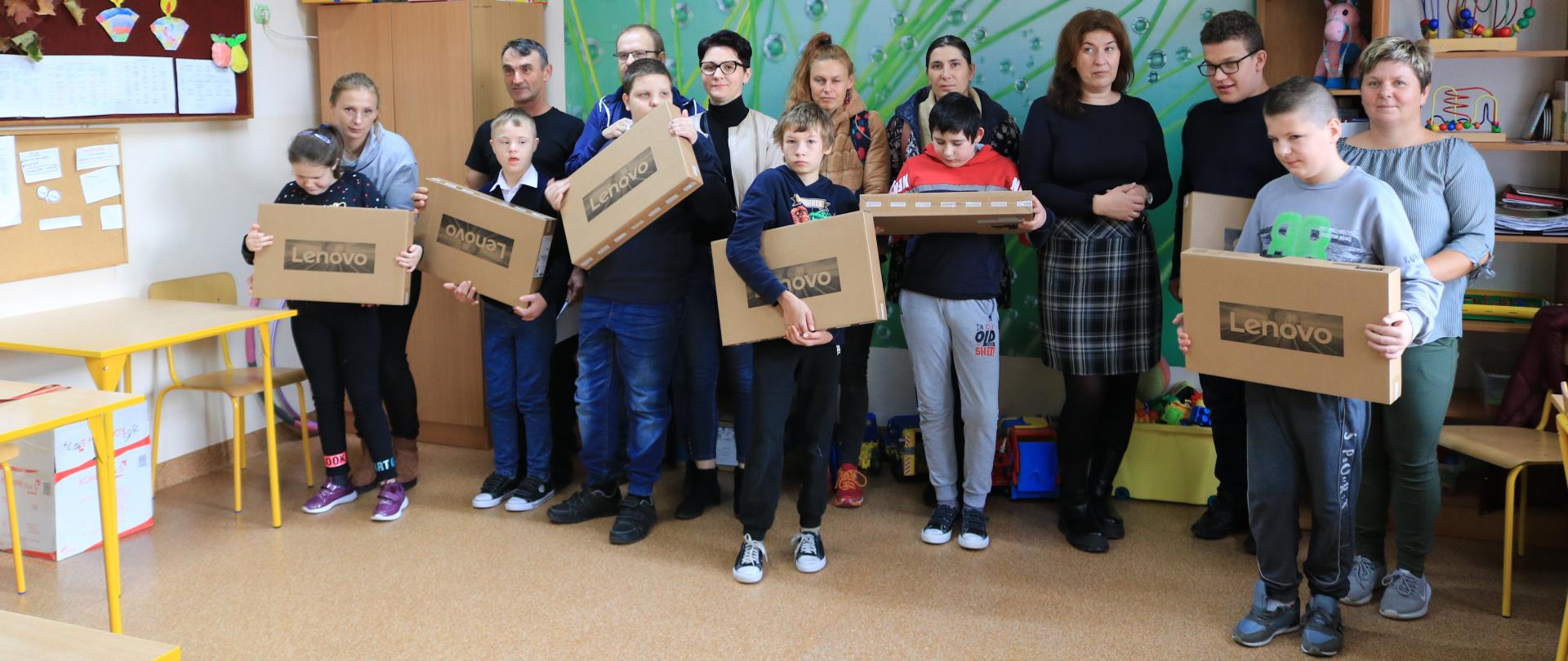 Zdjęcie przedstawia uczniów klasy IV Szkoły Podstawowej Specjalnej z otrzymanymi laptopami wraz z ich rodzicami i opiekunami. 
