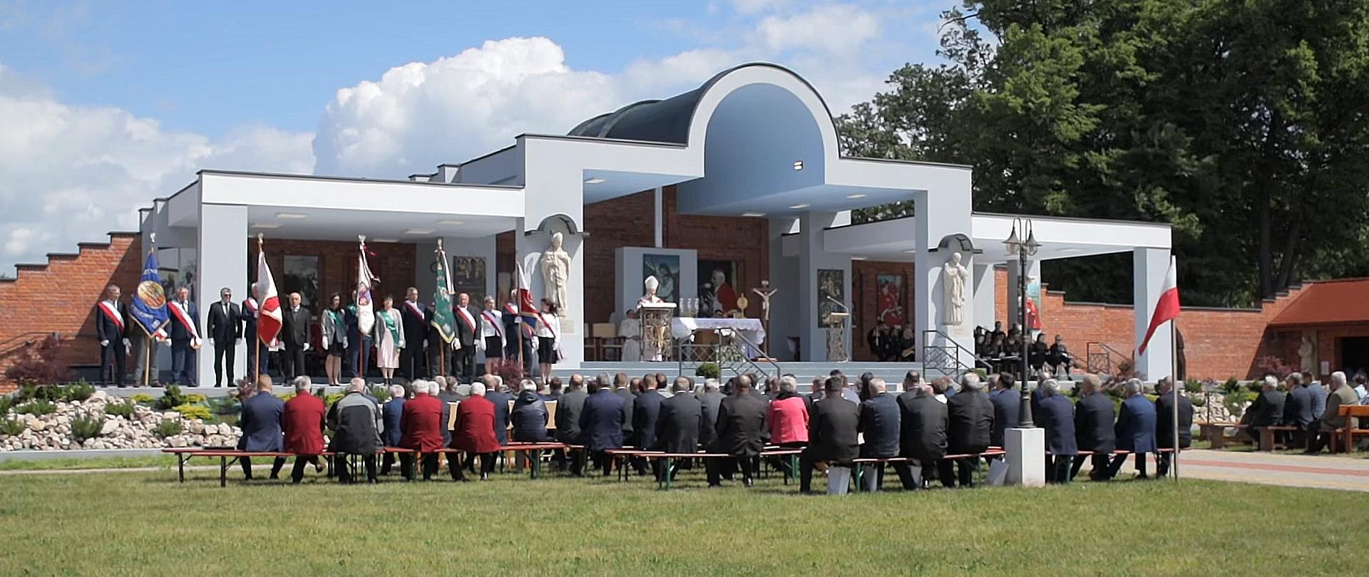 Kadr z filmu udostępnionego przez Telewizję Małopolską. Ołtarz polowy w szczepanowskim Panteonie Świętych i Błogosławionych. Na lewo od ołtarza stoją sołtysi z pocztami sztandarowymi. Przed ołtarzem siedzą sołtysi i zaproszeni goście. 