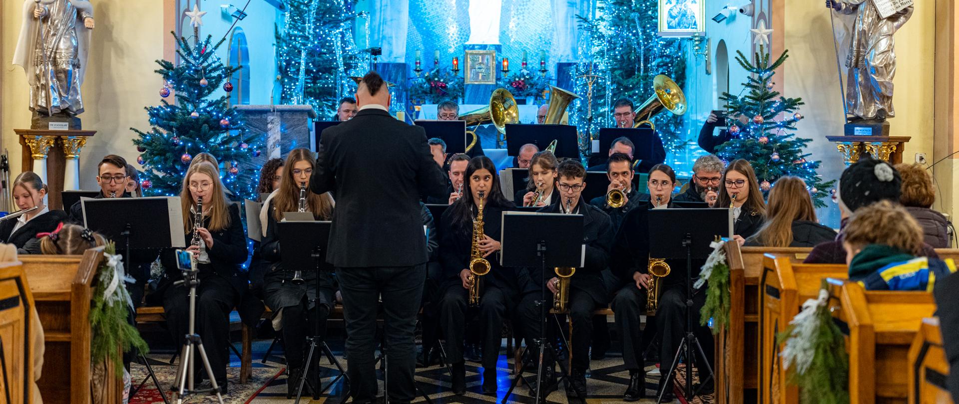 Parafialna orkiestra dęta podczas Koncertu Noworocznego w kościele w Leńczach – muzycy w czarnych strojach grają na instrumentach dętych, a dyrygent stoi przodem do zespołu. W tle ołtarz, świąteczne choinki z niebieskim oświetleniem oraz wierni siedzący w ławkach.
