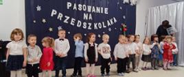 Przedszkolaki stoją w sali na tle ścianki "Pasowanie na przedszkolaka"