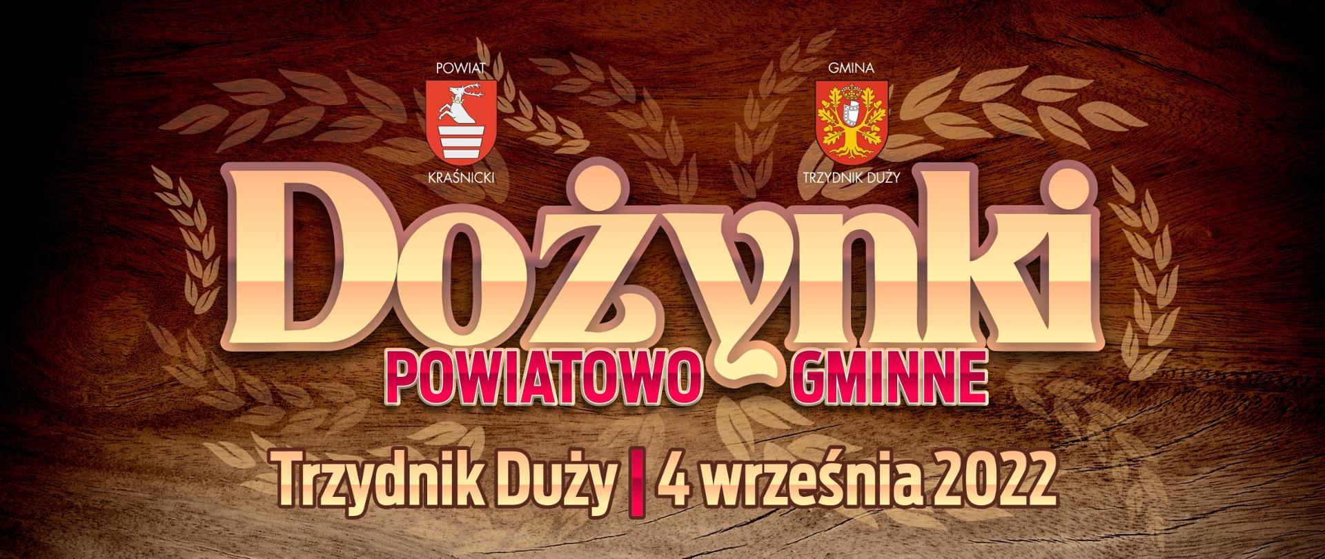 Grafika przedstawia stylizowany napis "Dożynki Powiatowo Gminne". Słowo Dożynki w odcieniach złota, poniżej czerwone słowa Powiatowo i Gminne. Nad napisem znajdują się herby: Powiatu Kraśnickiego i Gminy Trzydnik Duży. Poniżej znajduje się napis w odcieniach złota: Trzydnik Duży 4. września 2022. Tło stanowi zdjęcie drewnianej deski w odcieniach brązowych.