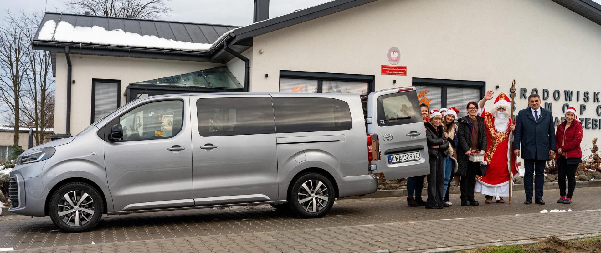 Grupa osób, w tym Święty Mikołaj w czerwonym stroju i czapce, stoi obok srebrnego busa zaparkowanego przed budynkiem Środowiskowego Domu Samopomocy. Uczestnicy uśmiechają się i pozują do zdjęcia, a część z nich ma na głowach mikołajowe czapki. W tle widoczna elewacja budynku z tablicą informacyjną oraz zimowa sceneria z resztkami śniegu na dachu i trawie.
