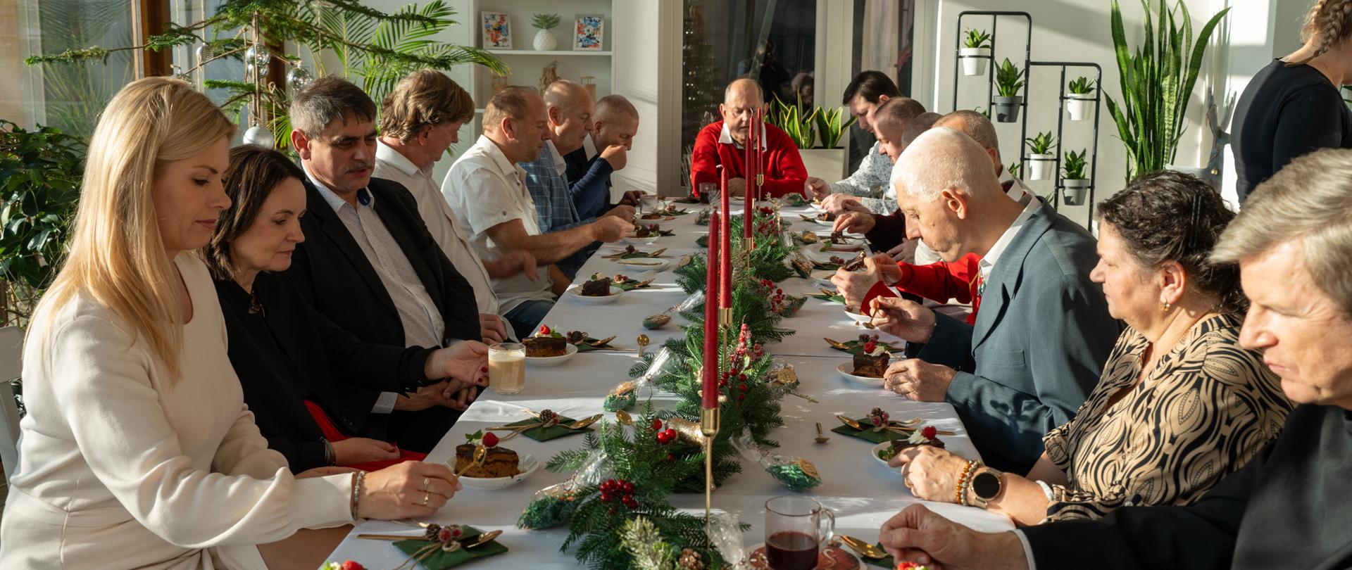 Uczestnicy spotkania świątecznego w Domu Pomocy Społecznej w Zebrzydowicach siedzą przy długim, odświętnie udekorowanym stole, wspólnie spożywając posiłek; na stole widoczne są świąteczne dekoracje z gałązek świerku i świec, a wnętrze sali jest jasne i pełne naturalnego światła.