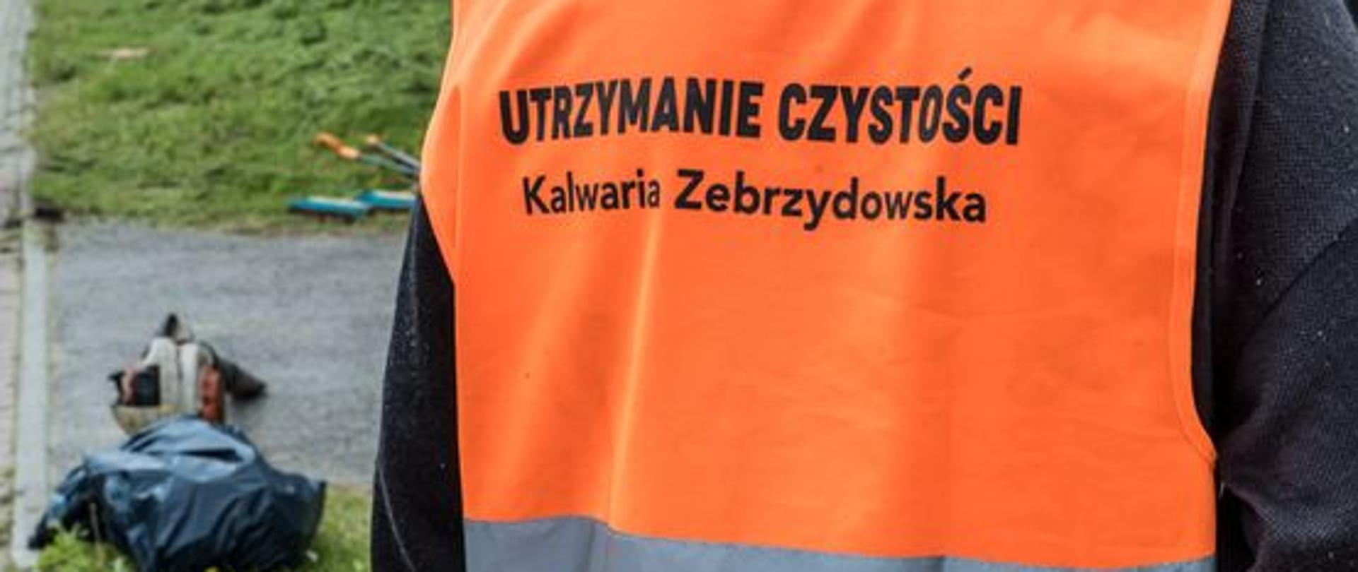 Plecy pracownika interwencyjnego noszącego pomarańczową kamizelkę odblaskową z napisem "Utrzymanie czystości Kalwaria Zebrzydowska". Po prawej stronie kadru dmuchawa do liści i czarny worek na śmieci.