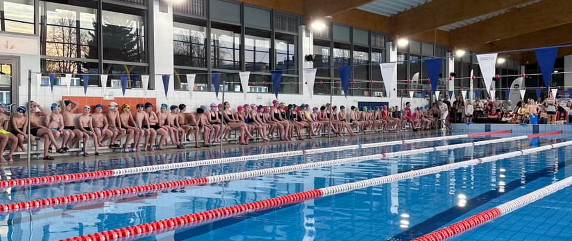 Zdjęcie przedstawia nieckę basenu a przy niej dzieci czekające na zawody.