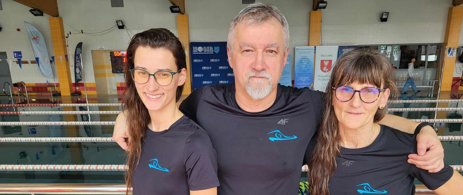 Na tle basenu w czarnych koszulkach z niebieskim logo Uczniowskiego Klubu Pływackiego "Fala" w Kraśniku stoją (od lewej) Zuzanna Mazurek-Frankowska, Grzegorz Mazurek i Iwona Mazurek. Grzegorz Mazurek.