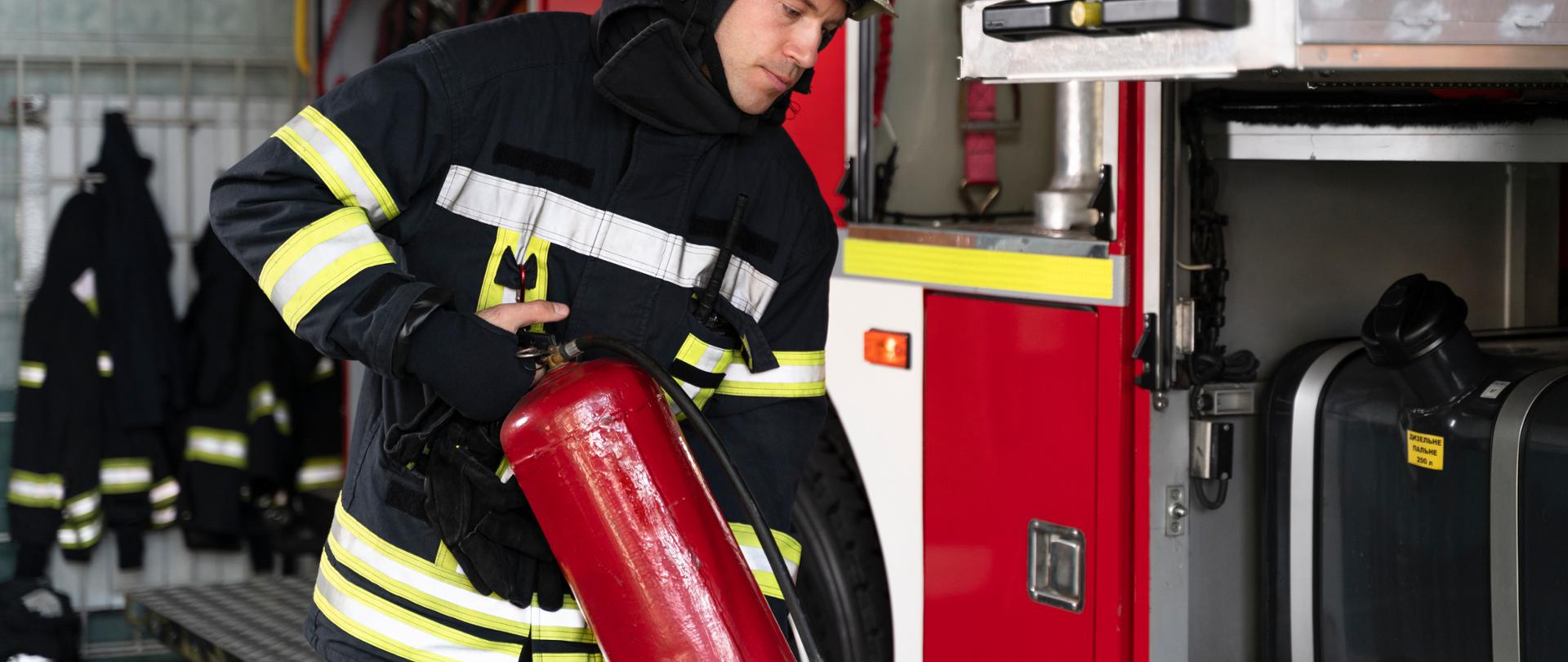 Strażak trzymający gaśnicę, w tle wóz strażacki.
