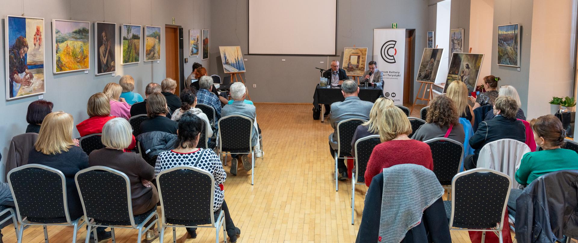 Na sali widowiskowej trwa spotkanie autorskie z udziałem publiczności. Uczestnicy siedzą w rzędach krzeseł, zwróceni w stronę dwóch prowadzących przy stoliku ustawionym na końcu sali. Na ścianach oraz na sztalugach wyeksponowano obrazy malarskie, a wnętrze sali wyróżniają drewniane belki stropowe i stonowana, galeryjna aranżacja.