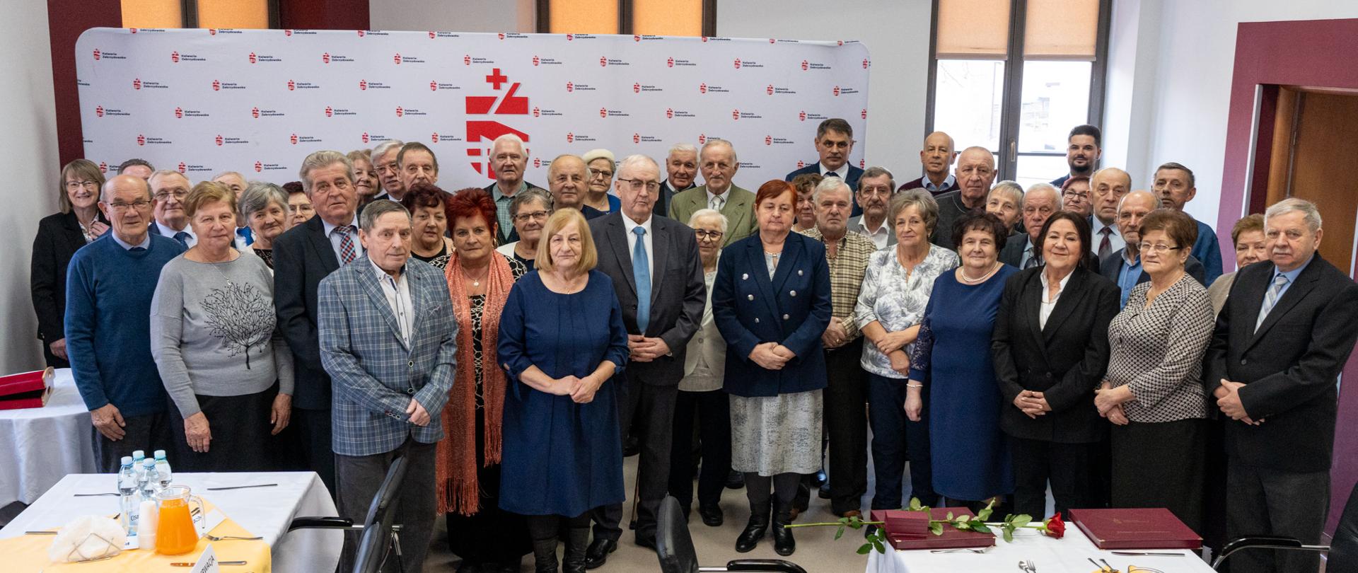 Grupowe zdjęcie kilkudziesięciu par i uczestników uroczystości jubileuszowej wykonane w sali spotkań. Na fotografii widoczne są osoby starsze, ubrane w eleganckie stroje, ustawione w kilku rzędach przed ścianką z herbem Kalwarii Zebrzydowskiej. W pierwszym planie stoją jubilaci, a na stole przed nimi leżą pamiątkowe dyplomy, pudełka z medalami oraz pojedyncza róża. W tle widać jasne wnętrze sali z oknami i elementami wystroju urzędowego.