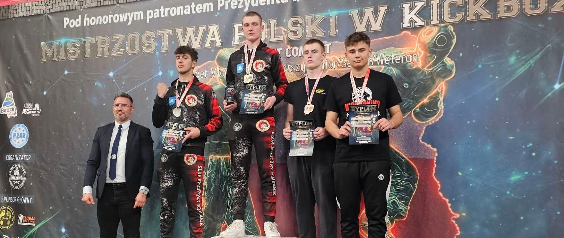 Najlepsi zawodnicy na podium zawodów. Szymon stoi na 3 stopniu wraz z innym zawodnikiem. Wszyscy wyróżnieni sportowcy mają zawieszone na szyjach medale, a w rękach trzymają dyplomy.