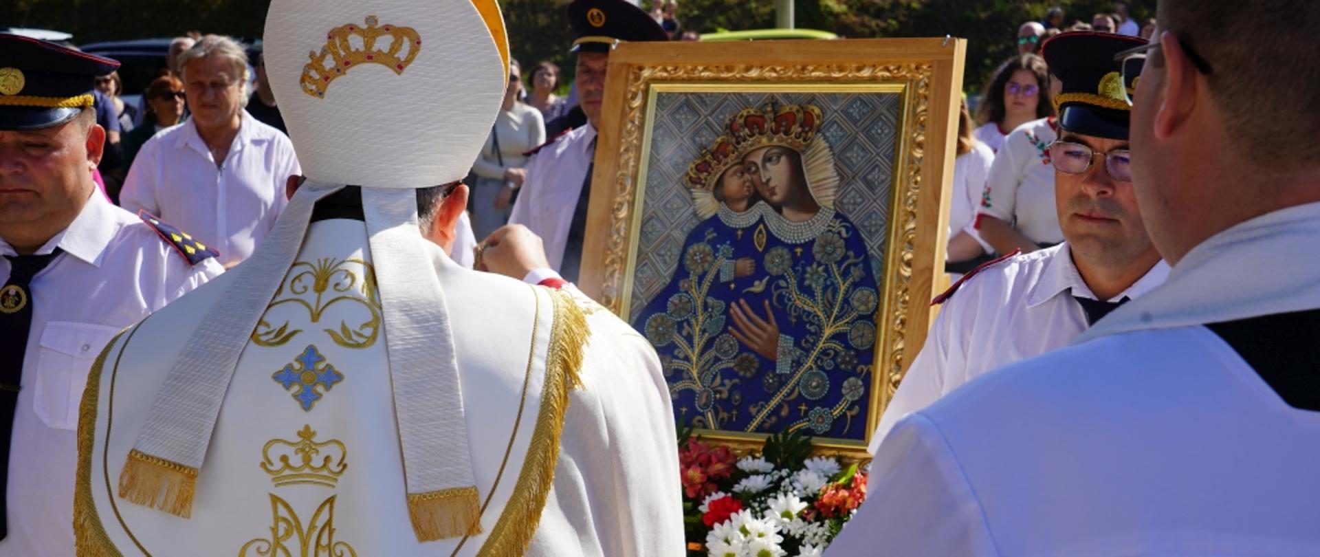 Uroczystość koronacji obrazu Matki Bożej – duchowny w szatach liturgicznych stoi przed ukoronowanym wizerunkiem Maryi z Dzieciątkiem, trzymanym przez strażaków podczas plenerowej ceremonii religijnej.