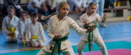 Układ karate wykonują dwie dziewczynki w tradycyjnym stroju kimono z zielonym pasem. W tle widać pozostałych uczestników turnieju
