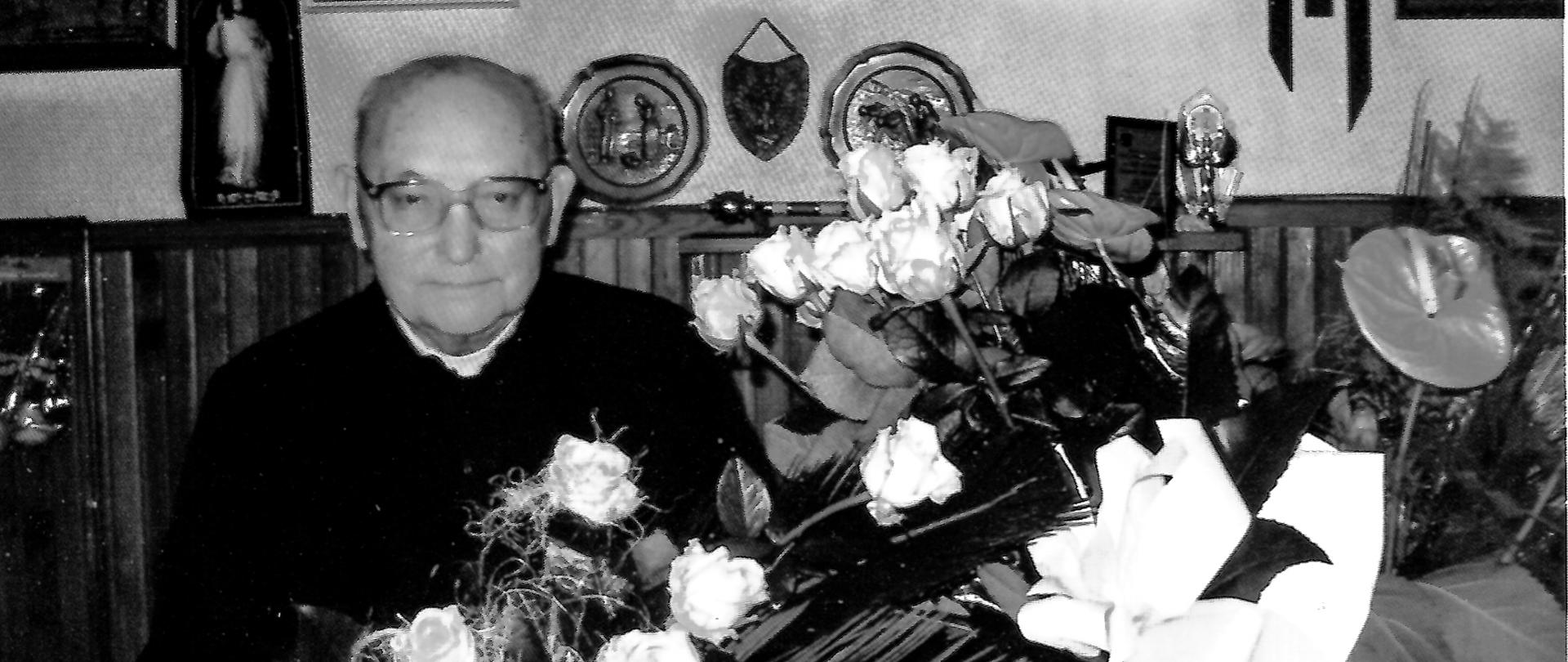 Czarno-białe zdjęcie przedstawiające starszego mężczyznę w sutannie siedzącego przy stole z bukietami kwiatów. W tle widoczne są obrazy i pamiątki religijne.