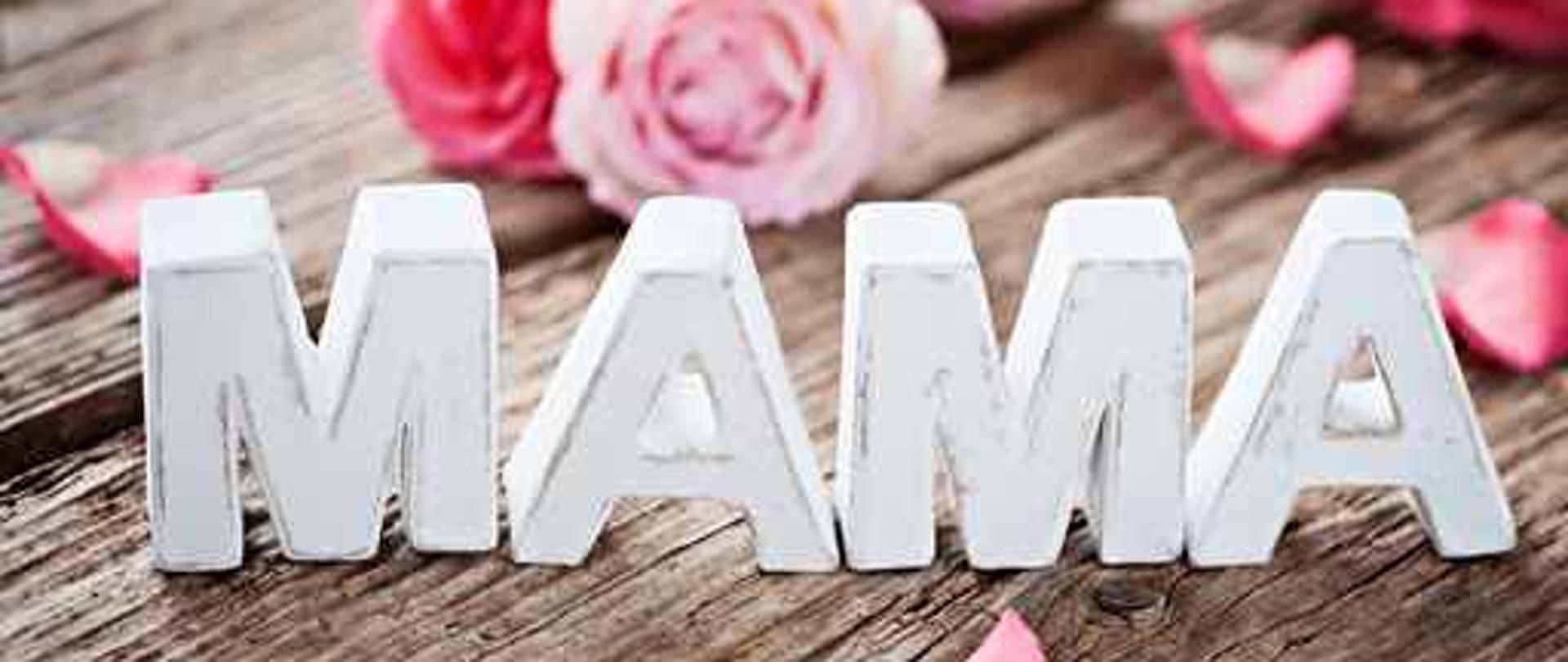 Na drewnianym podłożu stoją litery które tworzą napis Mama w tle widać kwiaty