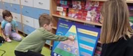 Dzieci z wykorzystaniem planszy edukacyjnej prezentują prowadzącej zajęcia jej poszczególne elementy