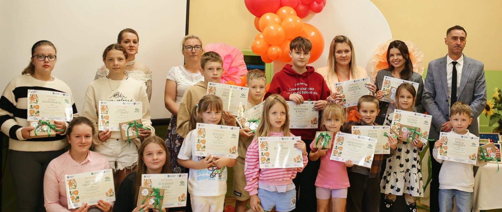 Grupa dzieci i dorosłych uczestników spotkania prezentuje dyplomy i nagrody; w tle znajduje się ekran i dekoracja z kolorowych balonów.