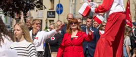Widać jak szczudlarz rozdaje uczestnikom obchodów 3 Maja flagi, uśmiechnięta kobieta w czerwonej marynarce i spodniach.