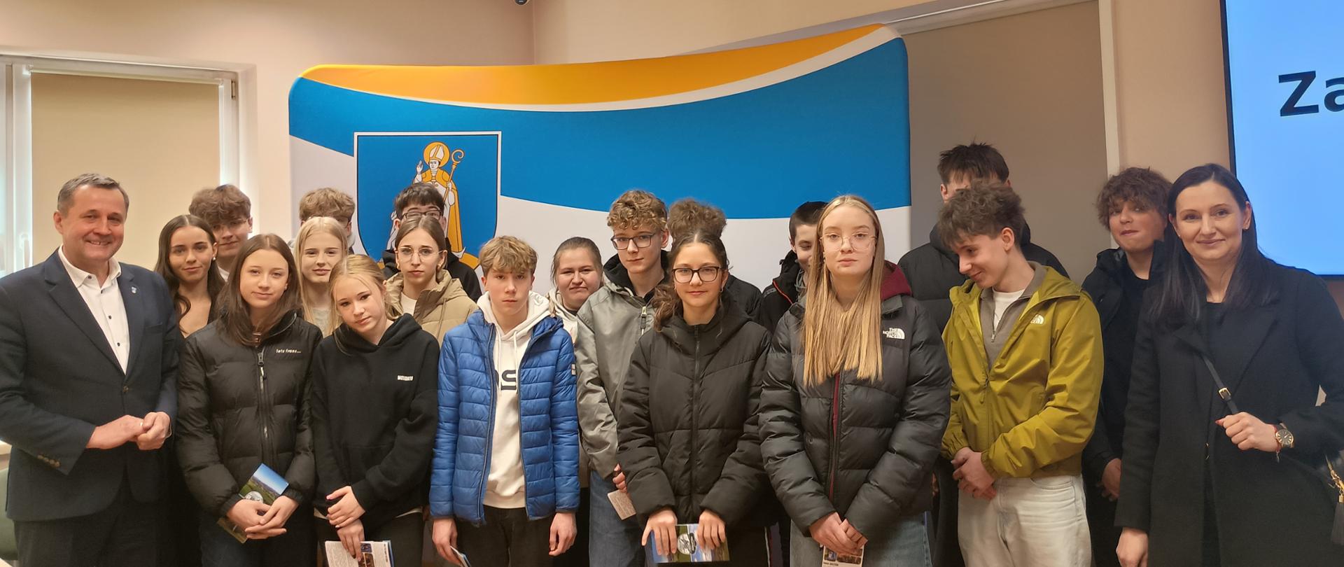 Grupa uczniów stojąca razem z dwojgiem dorosłych podczas wizyty w urzędzie. Młodzież ubrana w kurtki zimowe stoi przed ścianką z herbem gminy Raba Wyżna, a po lewej stronie stoi Wójt Gminy. W tle widać ekran prezentacyjny oraz wnętrze sali konferencyjnej.