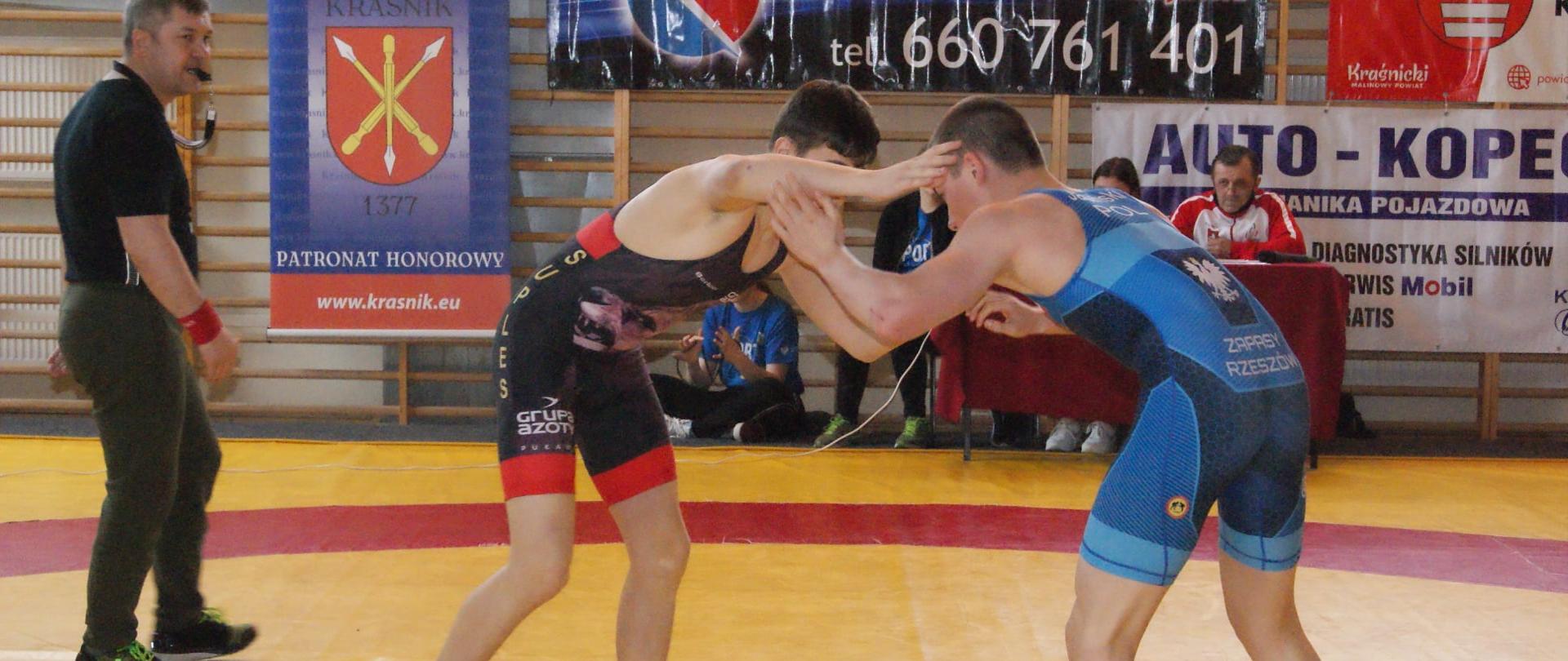Zdjęcie przedstawia dwóch zapaśników walczących na macie podczas Otwartych Mistrzostw Województwa Lubelskiego dzieci, młodzików i juniorów w zapasach – styl wolny, które odbywały się w dniu 8 maja 2021 r. w Zespole Szkół nr 3 w Kraśniku. Po lewej stronie z gwizdkiem w ustach stoi trener Suplesu Jarosław Kozak