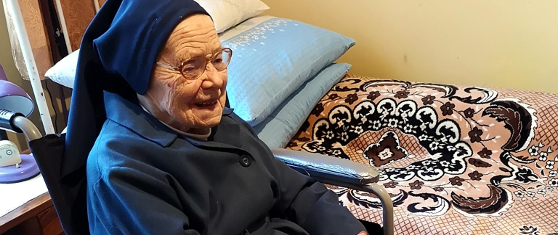 Siostra Szarytka siedzi uśmiechnięta na wózku, z tyłu nakryte kocem metalowe łózko i stos poduszek