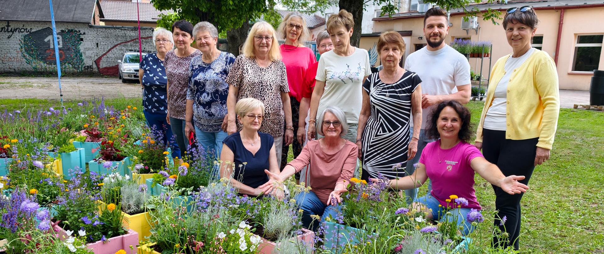 Grupa osób pozuje do zdjęcia na zewnątrz. Przed nimi ustawione są kolorowe skrzynki z kwitnącymi roślinami. Część osób stoi, a część kuca. W tle widoczne są drzewa, trawnik i budynki.